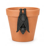 A black bat figurine hanging from the rim of a terracotta plant pot