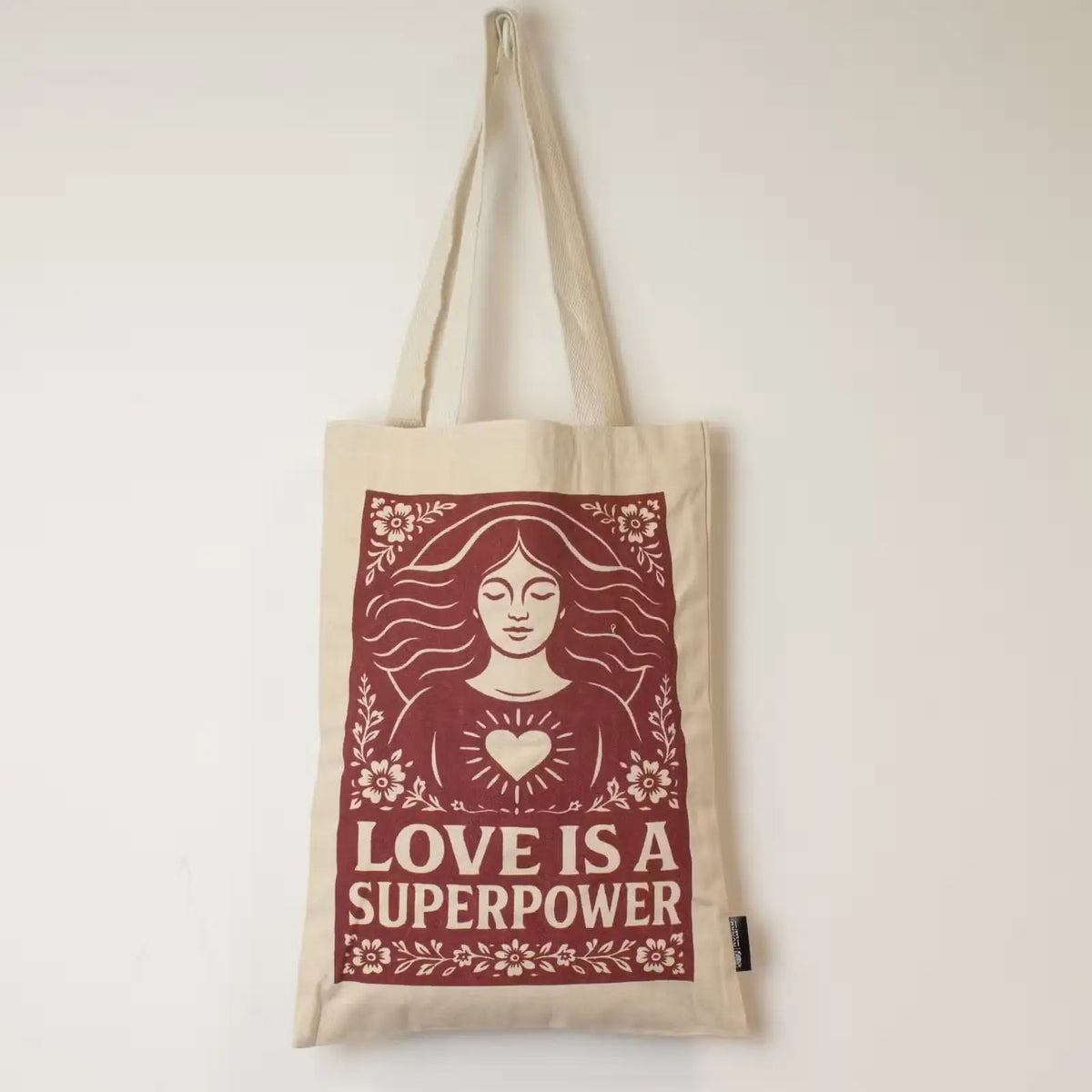 A natural cotton canvas tote bag featuring a large maroon graphic print of a woman with a heart and the text 'LOVE IS A SUPERPOWER'.