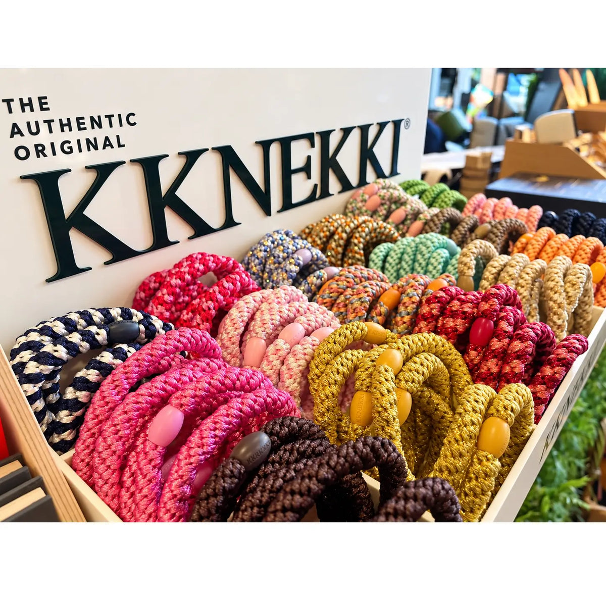 A display of colorful KKNEKKI braided rope hair ties arranged in a wooden tray, with the KKNEKKI logo visible on the display board behind them at Under the Sun Southend