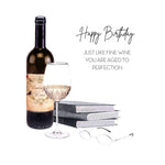 A watercolor-style birthday gift illustration featuring a dark wine bottle, a partially filled wine glass, stacked books, and eyeglasses, with the text 'Happy Birthday' and 'JUST LIKE FINE WINE YOU ARE AGED TO PERFECTION'.